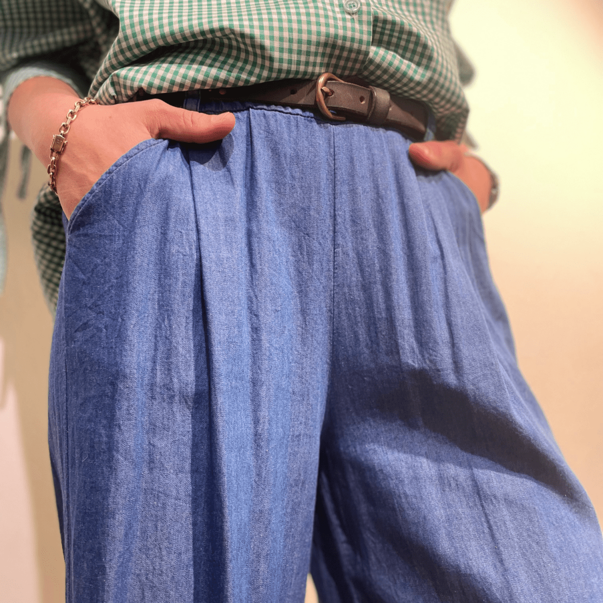 Et nærbillede af en person, der trækker i linningen på sine blå chambray bukser for at vise det løse fit. Personen har en grøn og hvid ternet skjorte stukket ned i bukserne og et brunt læderbælte med en guldspænde. Billedet fokuserer på personens maveområde fra lige under brystet til lårene.