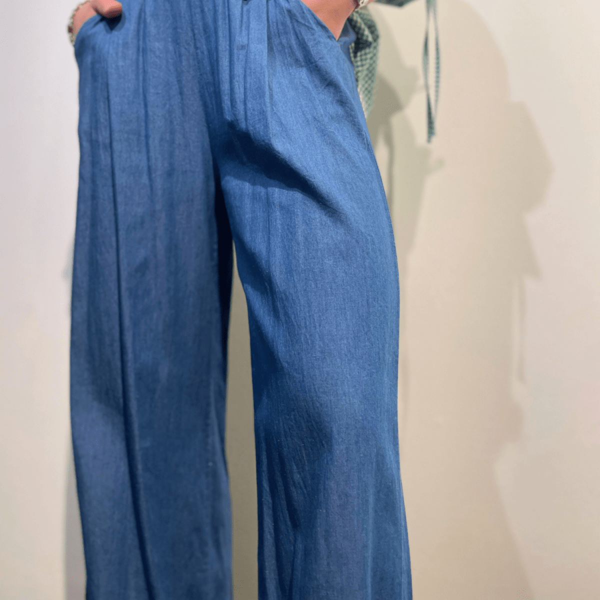  En person viser sideprofilen af blå chambray bukser med hånden på hoften. Skjorten, som er i et grønt og hvidt tern, er stoppet ind i buksernes linning. Dette mave-nærbillede er taget mod en neutral baggrund med en skygge af personen synlig på væggen til højre.